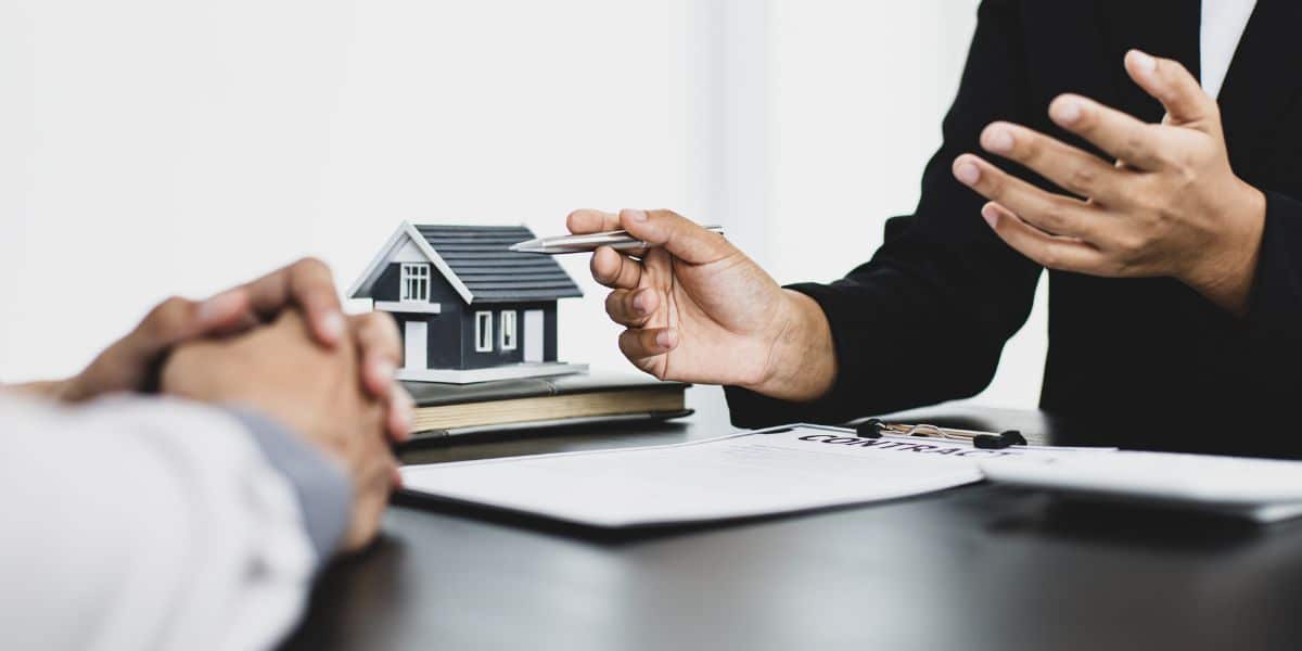 Close-up of a model house on a desk during a discussion between two people, with one gesturing while holding a pen